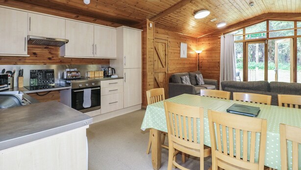 A cozy cabin-style kitchen with wooden cabinets, a dining area with a table and chairs, and a view of the surrounding forest through large windows. - Sycamore Lodge (Swadlincote)