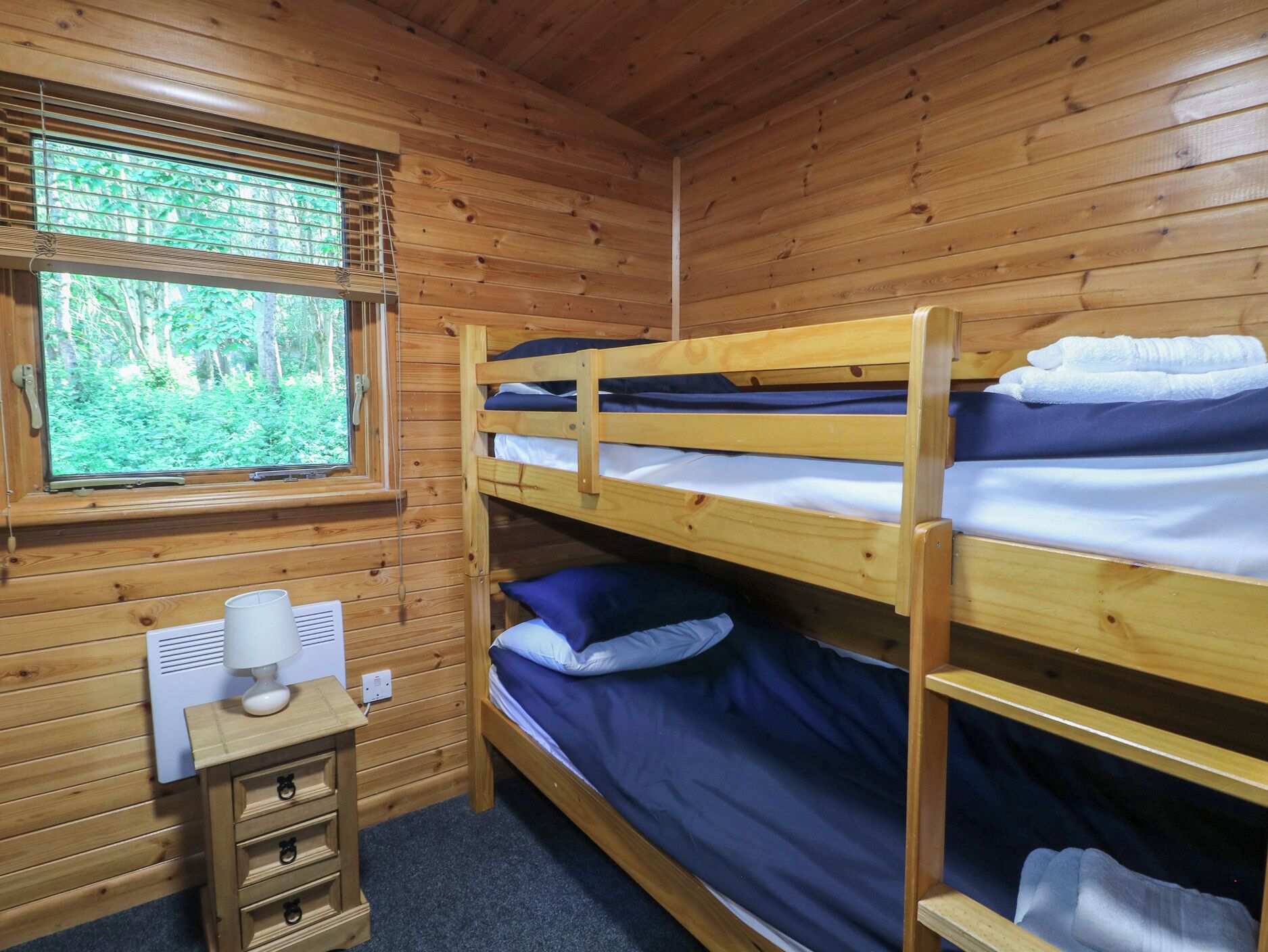 Habitación con literas, paredes de madera, ventana con persianas, mesita de noche pequeña con lámpara y toma de corriente.