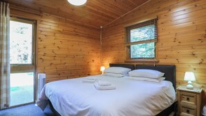 A wooden cabin bedroom with a bed, bedside lamps, and a window with blinds. - Cedar Lodge (Swadlincote)