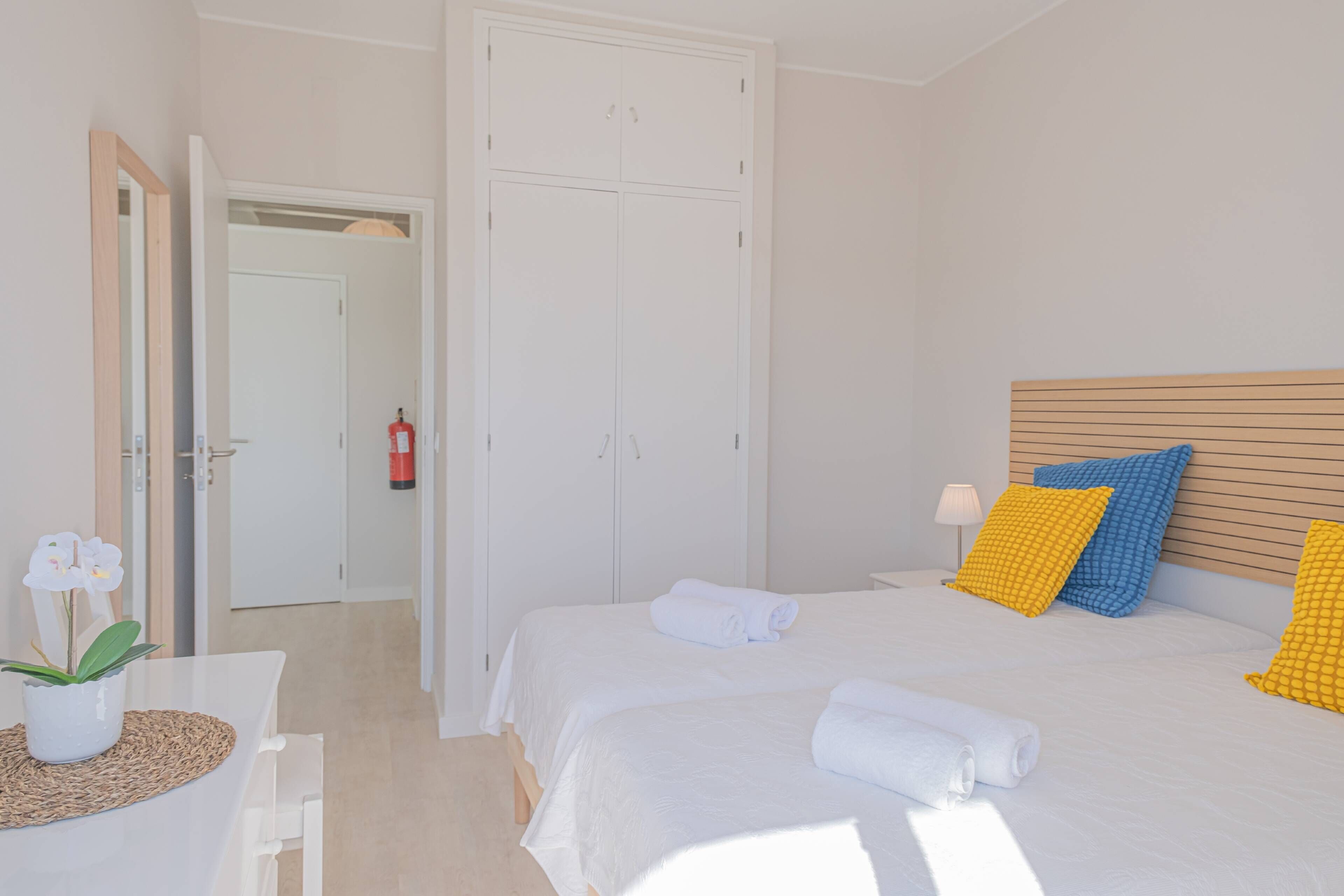A hotel room with a bed, a bedside table with a vase of flowers, a wardrobe, and a door leading to another room.
