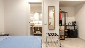 A hotel room with a bed, a small table, a bench, a mirror, a shelf with a red bag, and a coffee machine. - Ai Vicoli Ciambra's Alley - Suite Guglielmo by Wonderful Italy (Monreale)