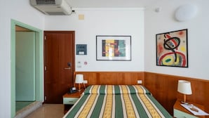 A hotel room with a bed, a bedside table, a lamp, a wall-mounted air conditioner, and a door. - Relaxing stay in a cozy double room in Bibione (Bibione)