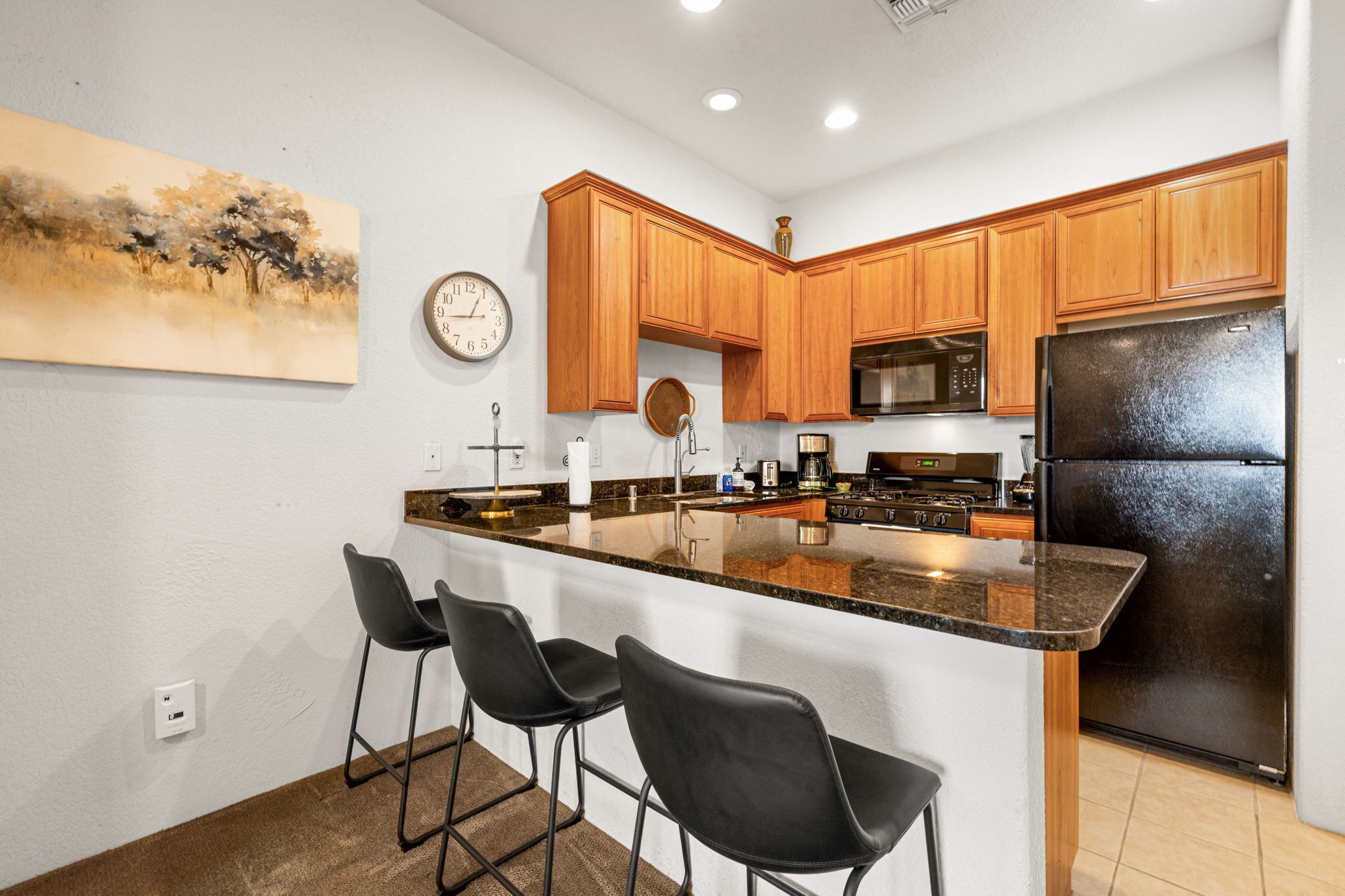 Cocina moderna con armarios de madera, un refrigerador negro, un microondas y un lavabo. Hay tres taburetes negros y un cuadro en la pared.