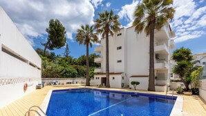 A swimming pool with a tiled deck, surrounded by palm trees and a white building with balconies.