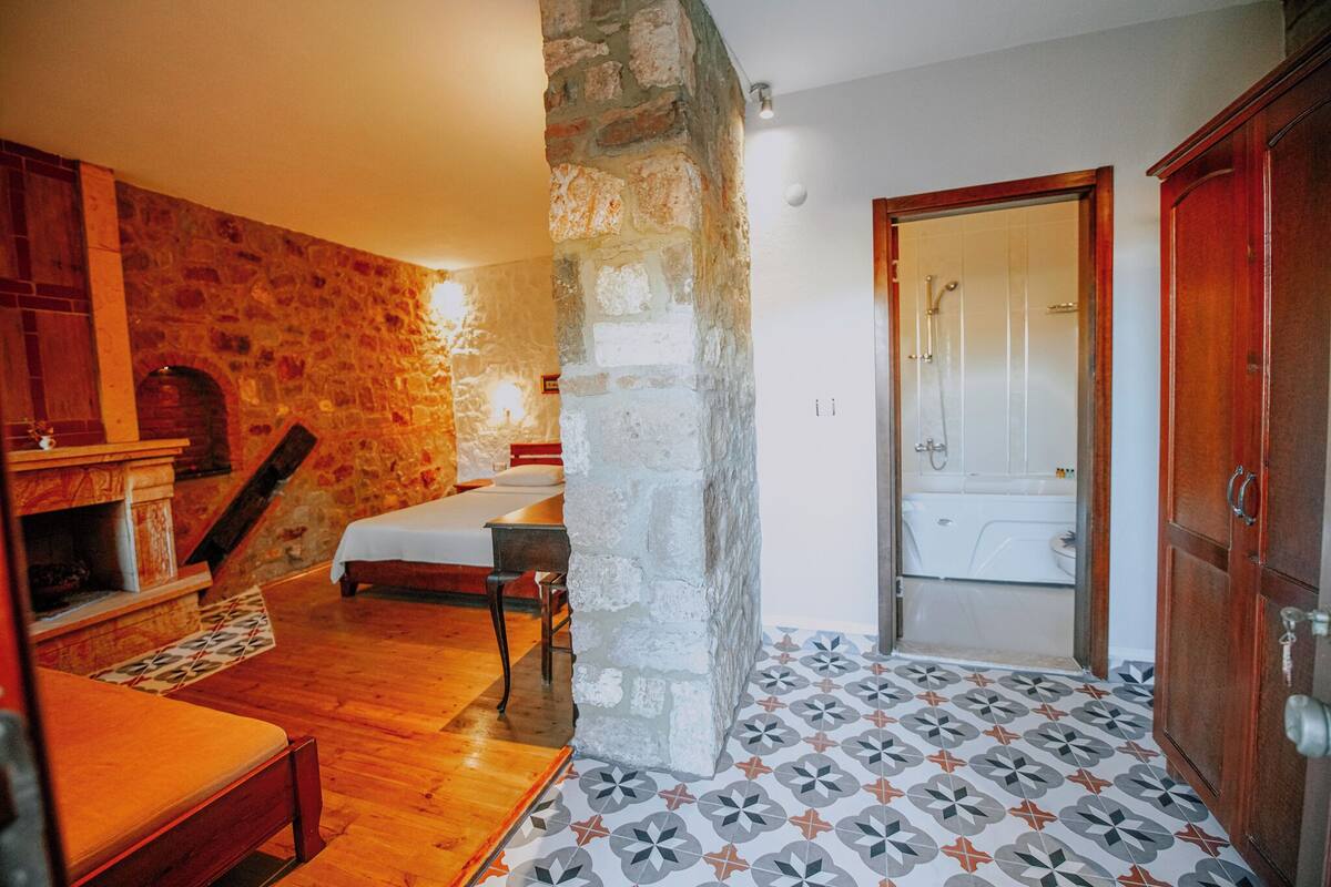 A room with a stone wall, a bed, a wooden bench, a tiled floor, and a bathroom with a shower.