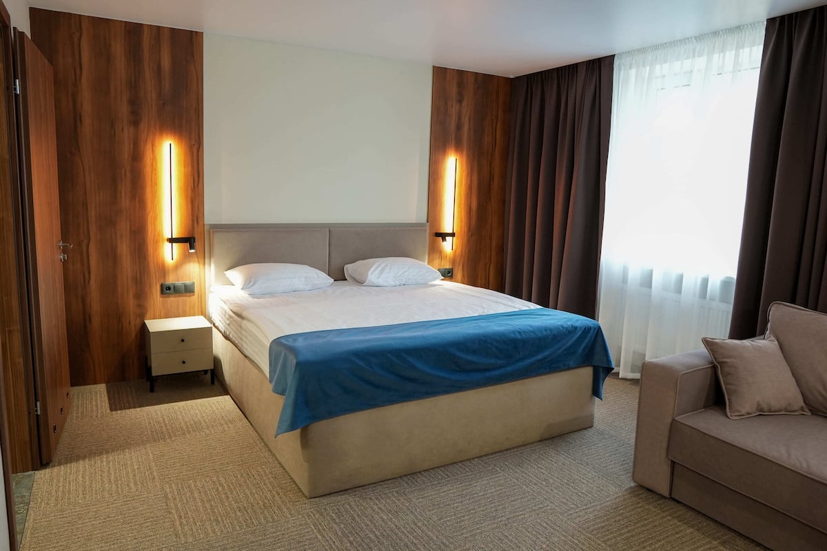 Quarto de hotel com uma cama grande, duas mesas de cabeceira, um criado-mudo, um sofá e uma janela com cortinas.