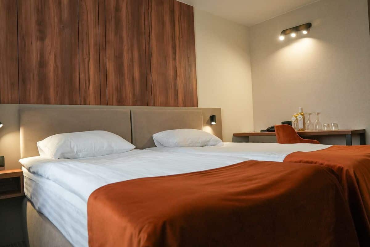 Quarto de hotel com uma cama grande, cabeceira de madeira, mesa de cabeceira e escrivaninha com luminária e garrafa.