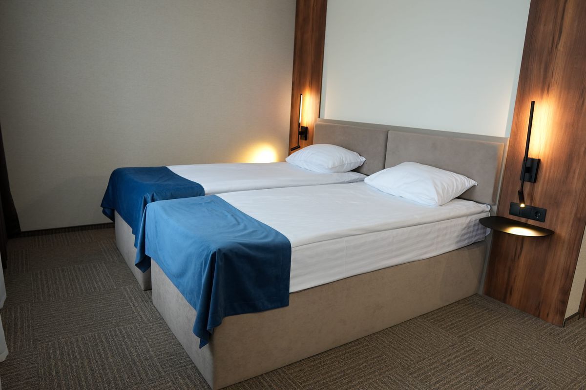 Habitación de hotel con dos camas, cada una con una colcha azul y almohadas blancas. Hay luces fijadas en la pared a ambos lados de las camas.