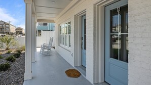 Fachada de ladrillo blanco con puerta azul, un porche cubierto y un pequeño jardín con una mecedora blanca y un tapete tejido.