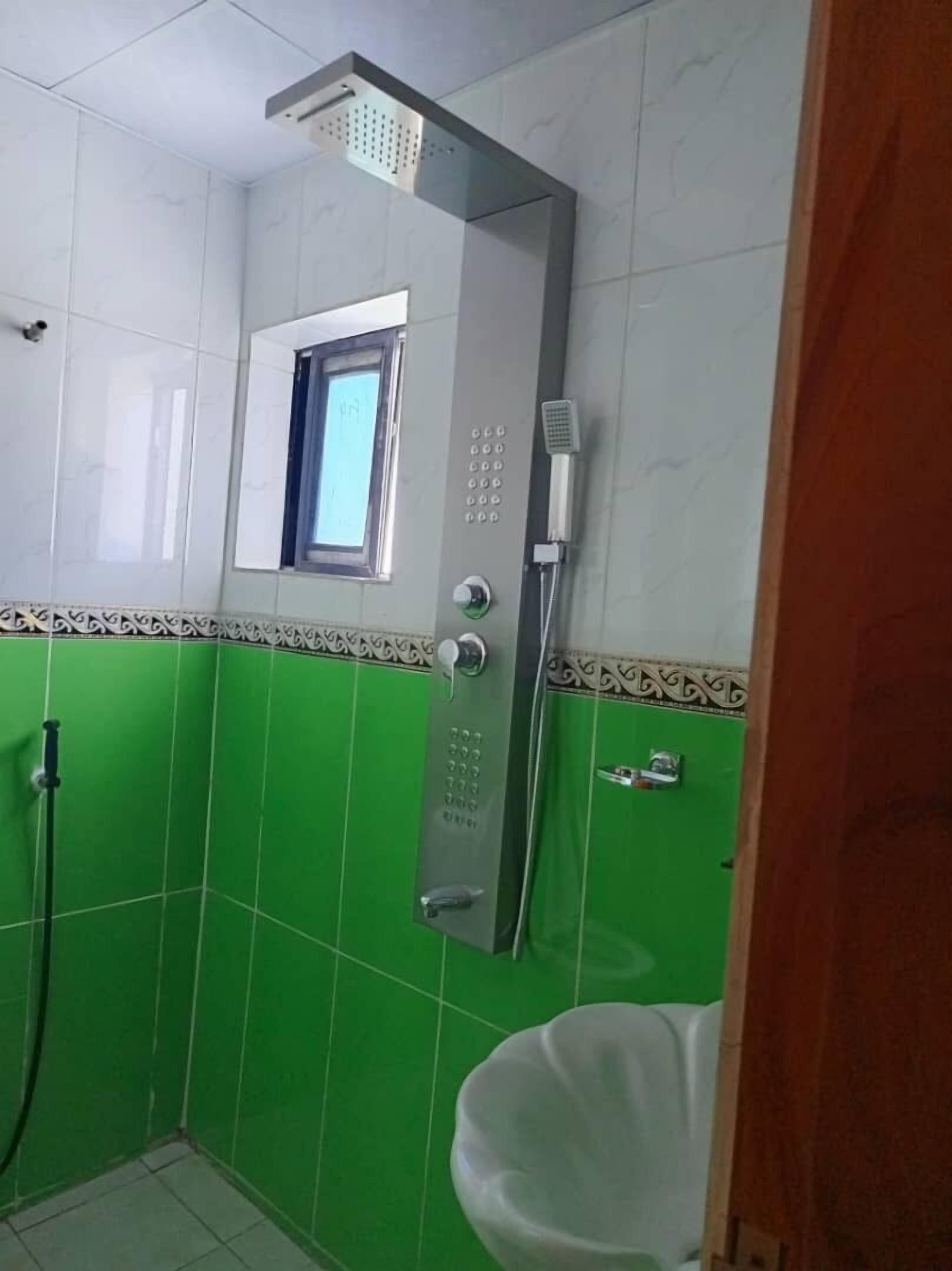 Ein Badezimmer mit grünen Fliesen, einer Dusche mit Bedienfeld und einem Fenster.