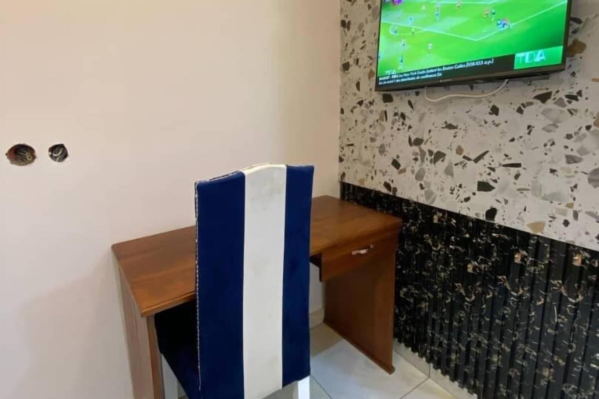 A room with a television mounted on a speckled wall, a wooden desk, and a chair with a blue and white striped backrest.