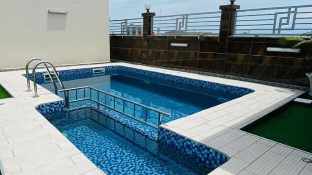 A swimming pool with a tiled deck, a satellite dish on the building, and a clear sky. - Guiko Palace Hotel  (Douala)
