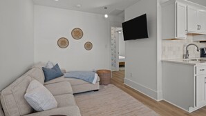 Ein modernes Wohnzimmer mit einem beigefarbenen Sofa, einem flachbildfernseher an der Wand und einer Küchenzeile mit weißen Schränken und einem Spülbecken.