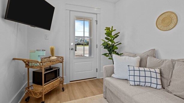 A modern living room with a flat-screen TV mounted on the wall, a wicker TV stand with a microwave, a beige sofa with patterned cushions, and a large window with a view of a building outside.