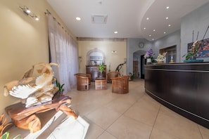 A hotel lobby with a reception desk, wooden chairs, a decorative sculpture, and a tiled floor. - Hotel Al Ritrovo (Castelluzzo)