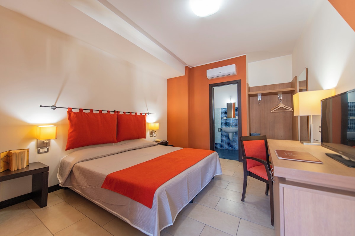 Habitación de hotel con cama, lámparas de noche, escritorio con silla, espejo y aire acondicionado.