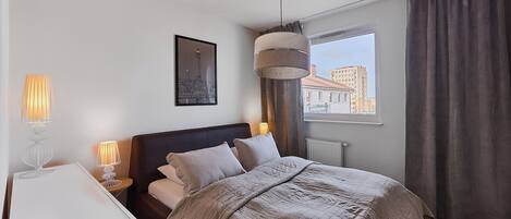 Un dormitorio con cama, lámparas de noche, un cuadro enmarcado, una ventana y cortinas.