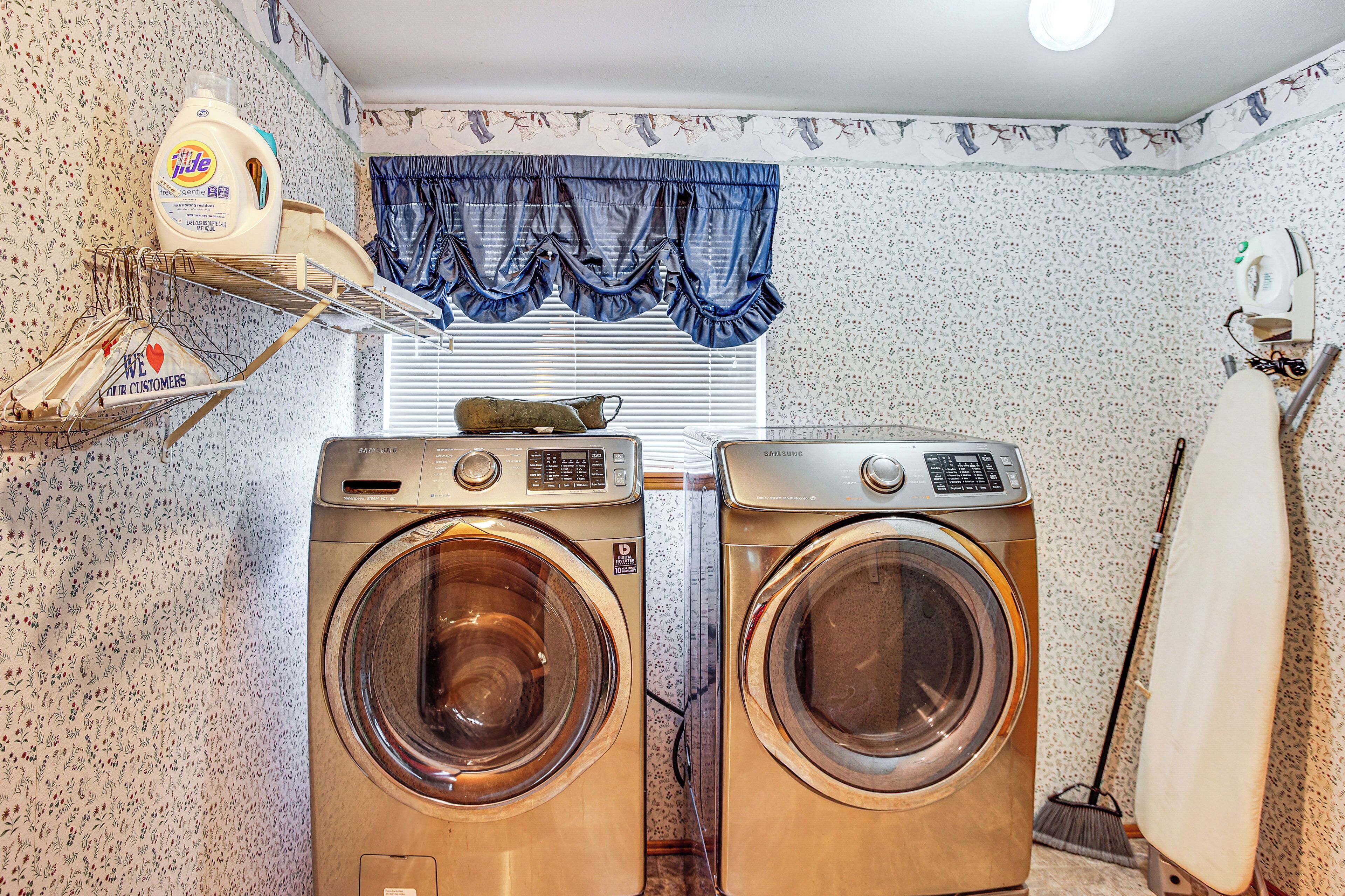 A laundry area with two front-loading washing machines, a drying rack, a bottle of detergent, and a window with blinds.