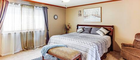 Un dormitorio con cama, una silla, un tapete, ventilador de techo y ventana con cortinas.