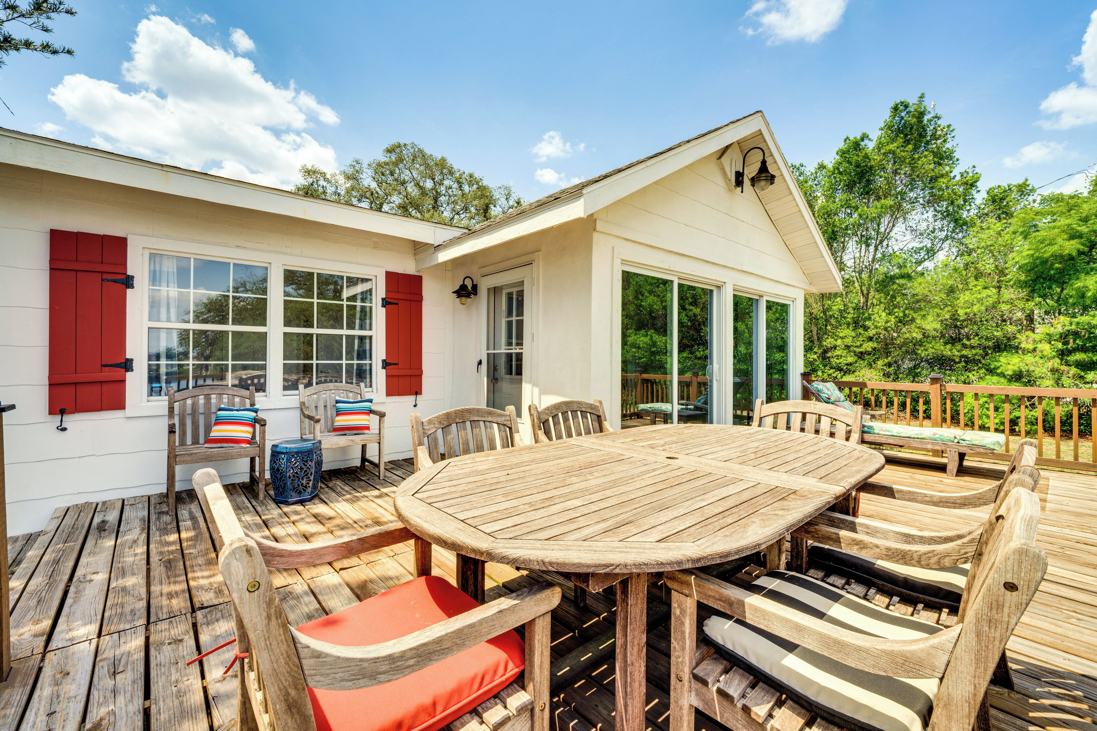 A wooden deck with a round table and chairs, a small porch with a bench, and a white building with red shutters.