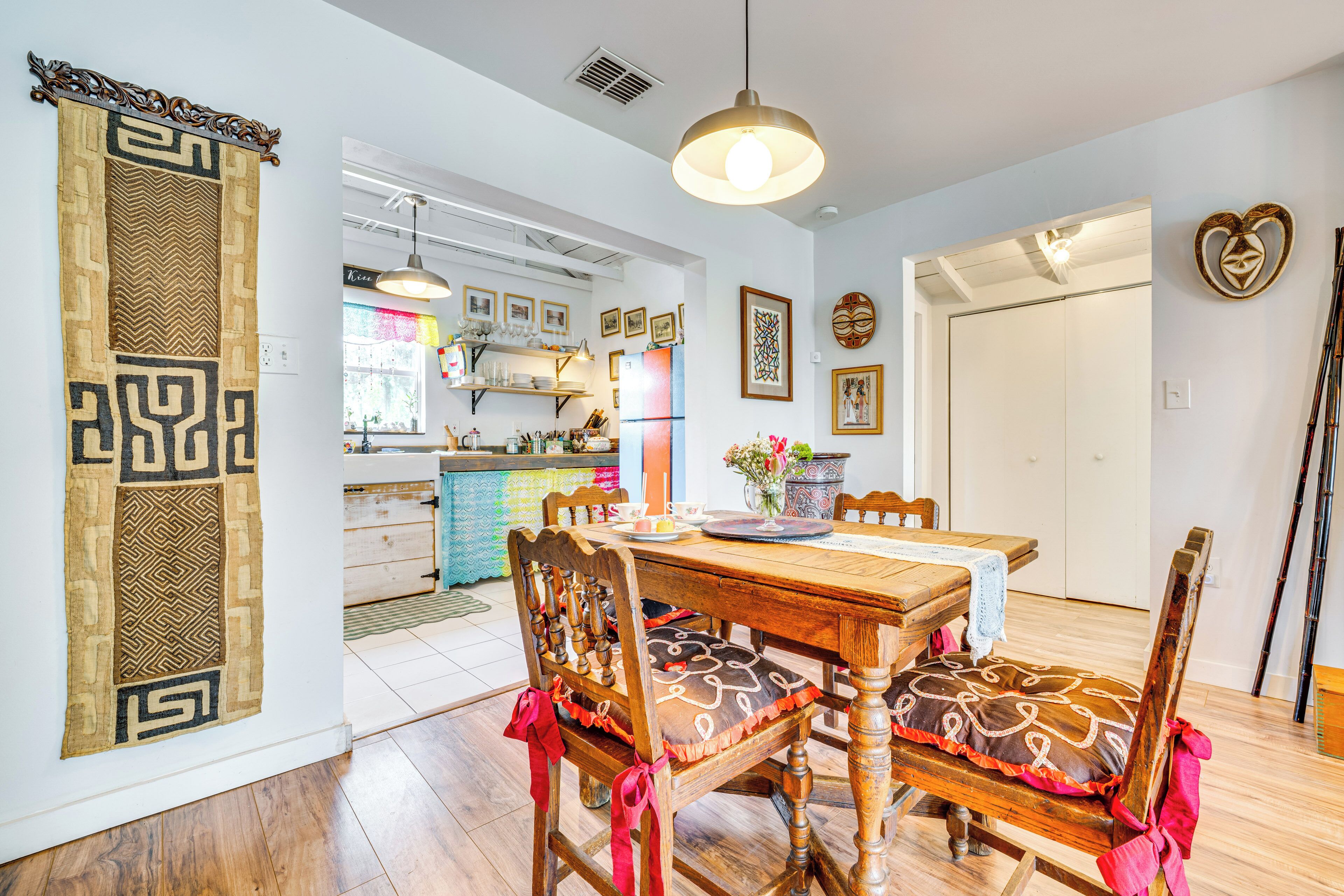 A dining area with a wooden table and chairs, a hanging light fixture, and a kitchen in the background.