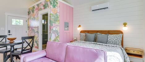Un dormitorio con cama, sofá rosa, mesa de comedor con sillas y una pared decorada con un mural floral.