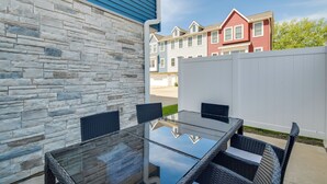 Un patio con una mesa de vidrio y sillas de mimbre, pared de piedra y vista a casas vecinas.