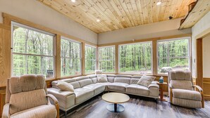 A spacious room with a large sectional sofa, two armchairs, and a round coffee table. The room features large windows offering a view of a forested area.