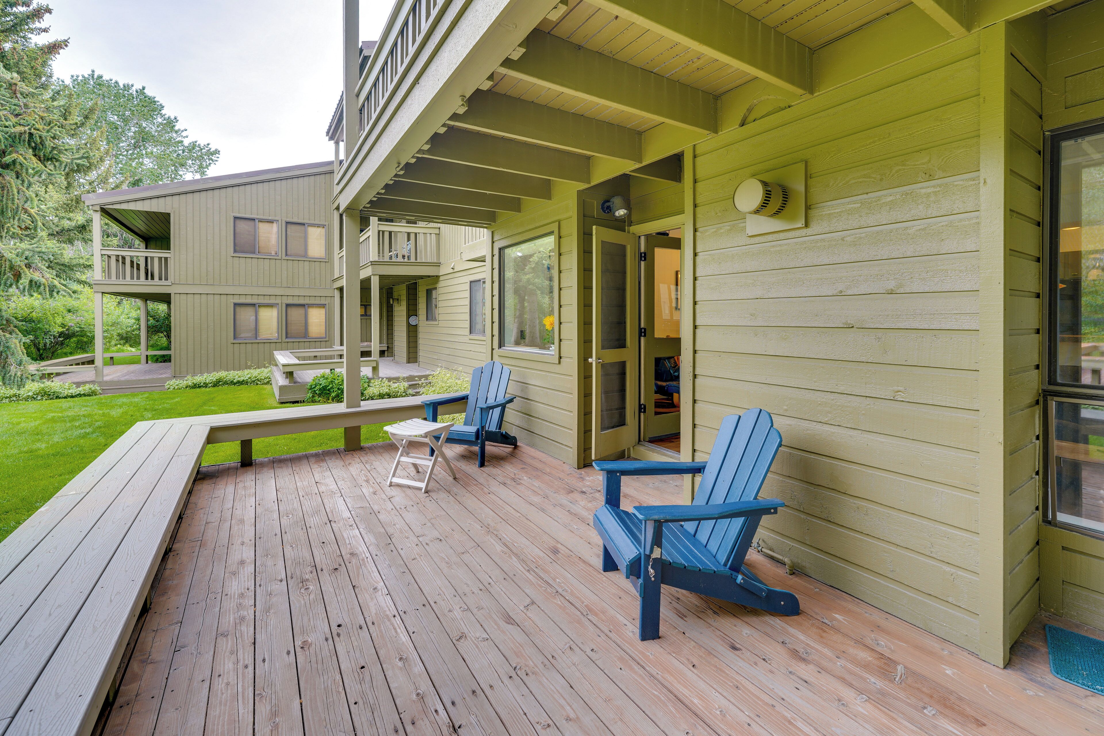 Une terrasse en bois avec deux chaises Adirondack bleues, un banc et un bâtiment doté d’un balcon.