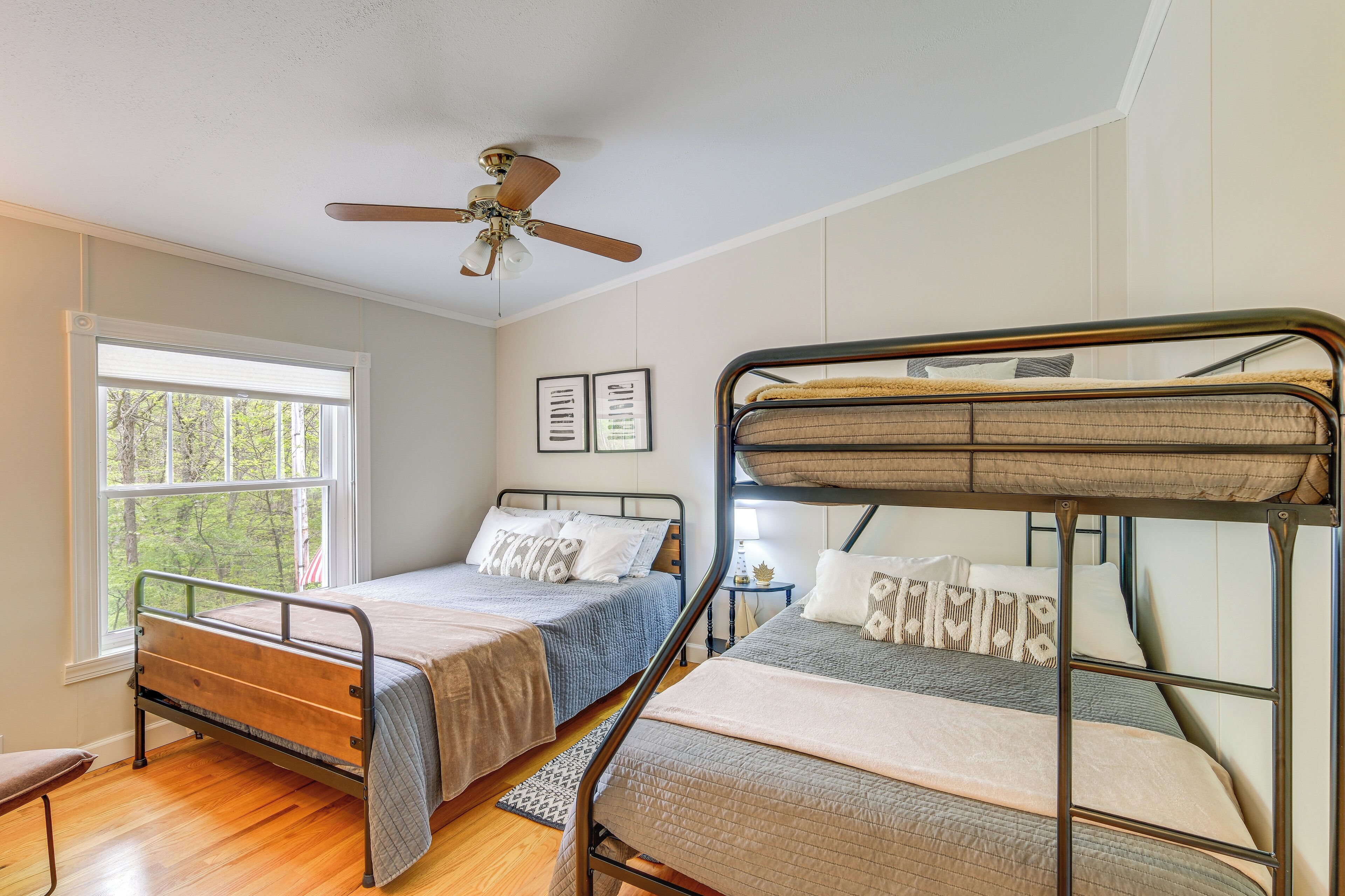 A room with two bunk beds, a ceiling fan, a window with blinds, and framed pictures on the wall.