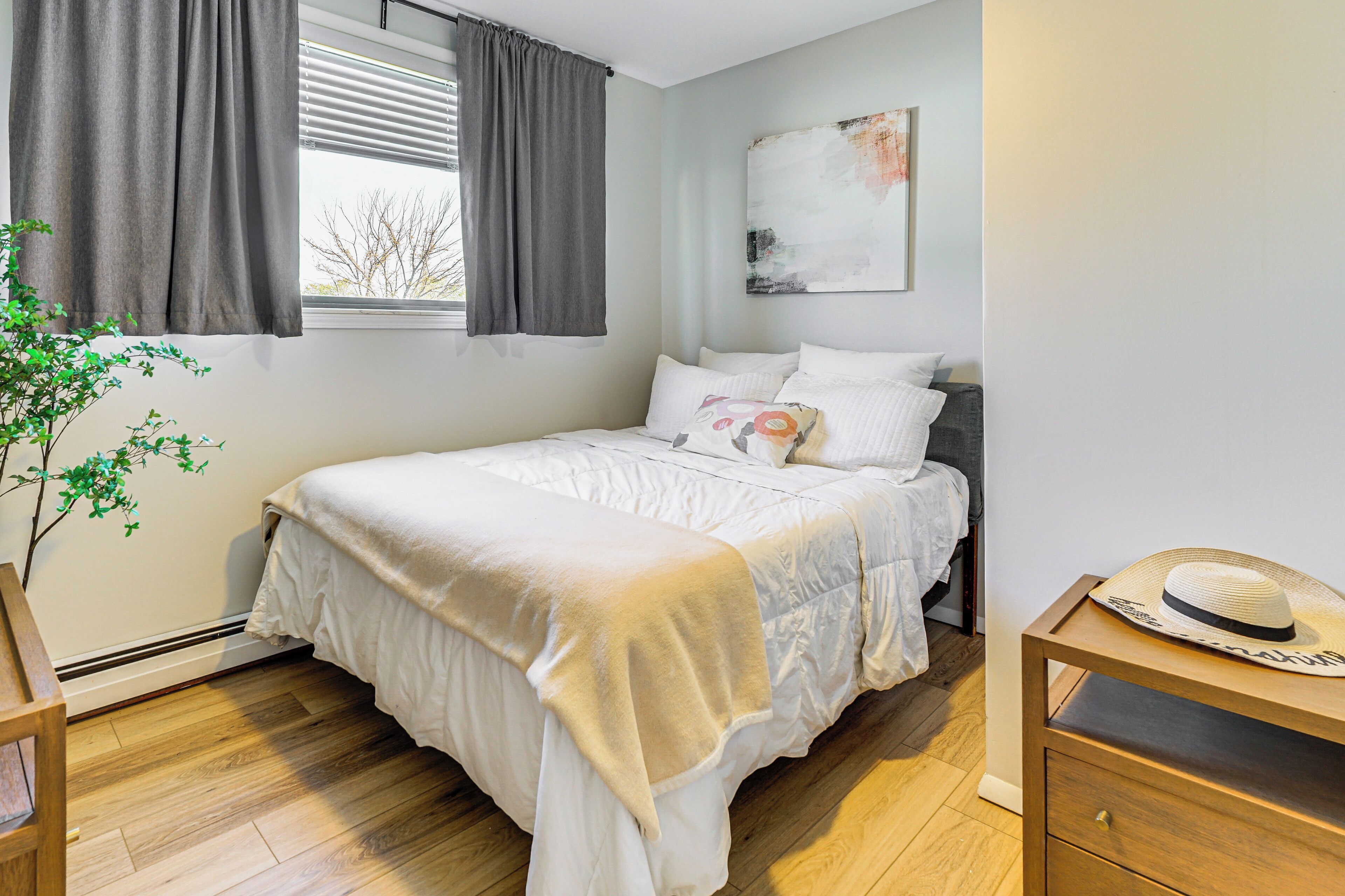 Une chambre à coucher bien rangée, avec un lit, une table de chevet, une œuvre d’art accrochée au mur et une fenêtre avec des rideaux.