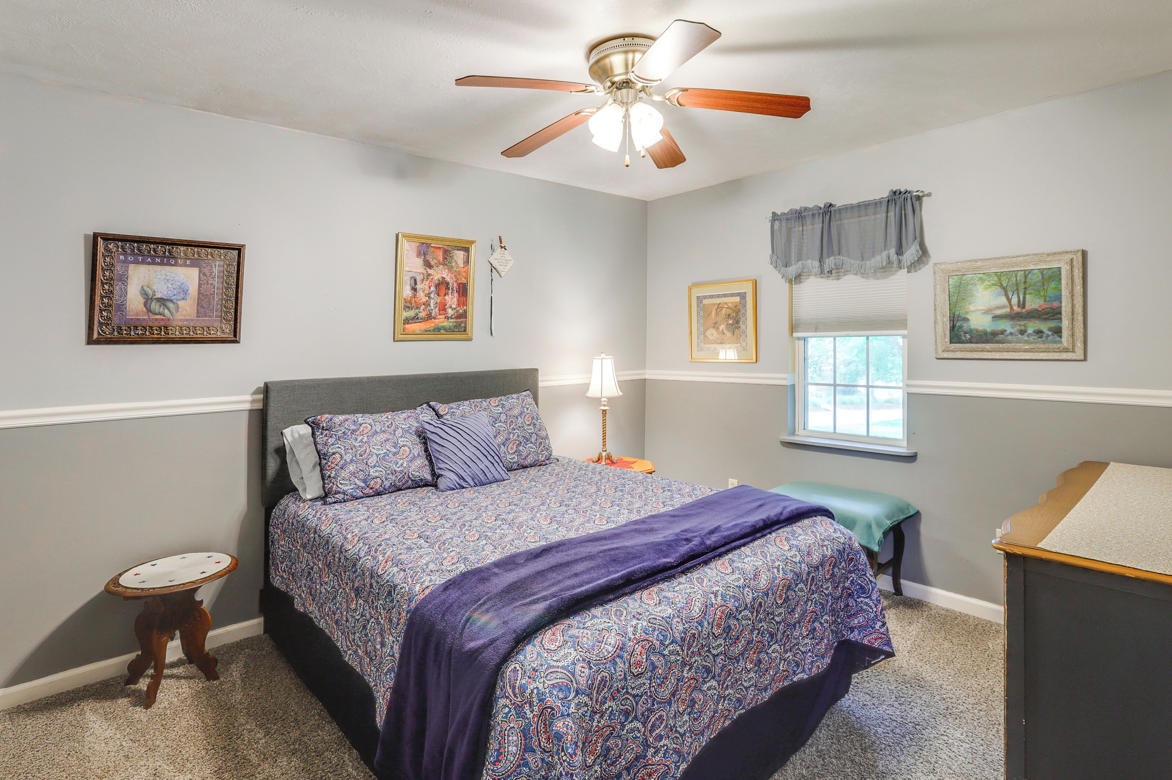 A bedroom with a bed, a ceiling fan, a bedside table, a lamp, and framed artwork on the walls.