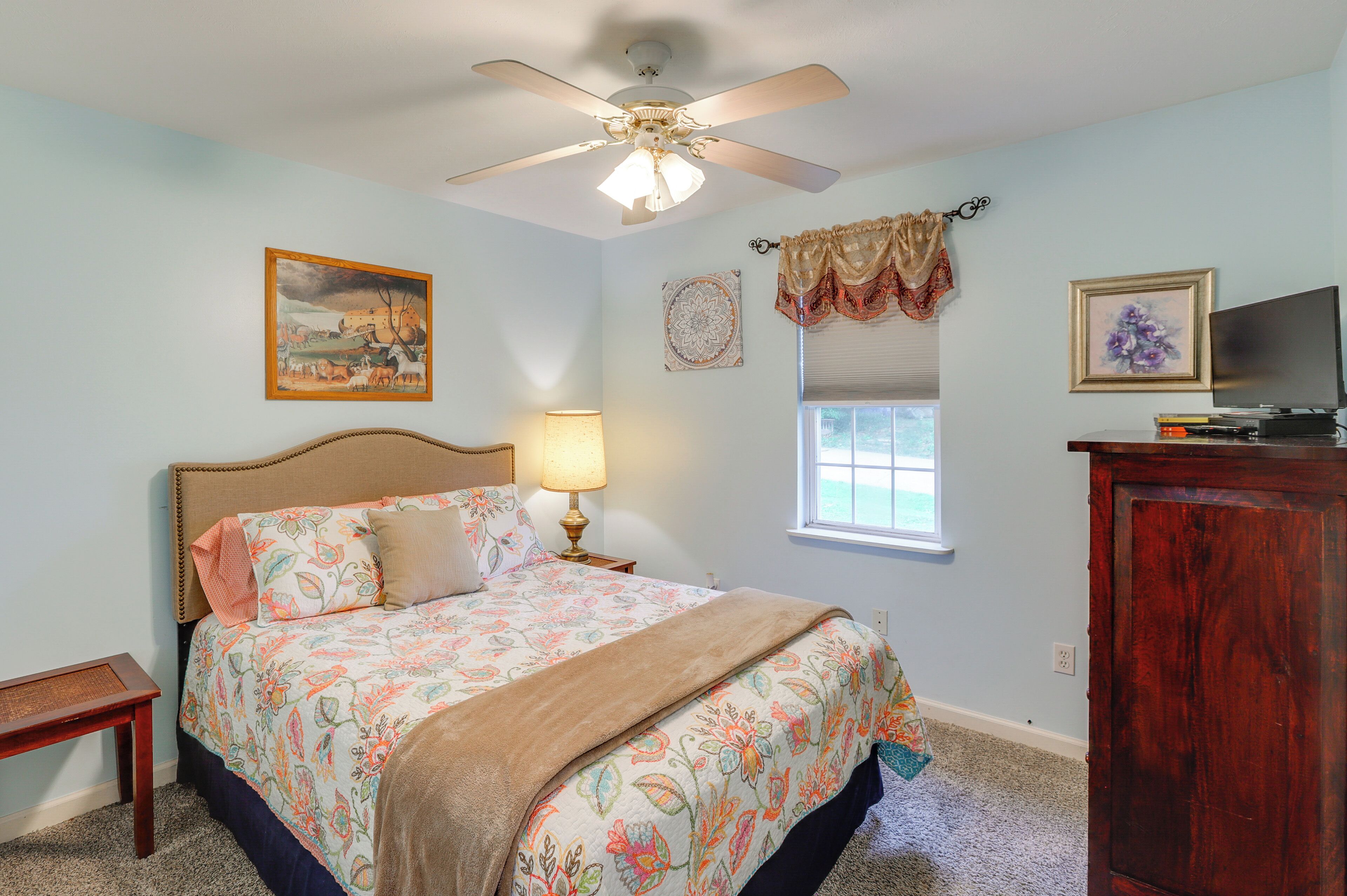 A bedroom with a bed, a ceiling fan, a window with blinds, a TV, and a painting on the wall.