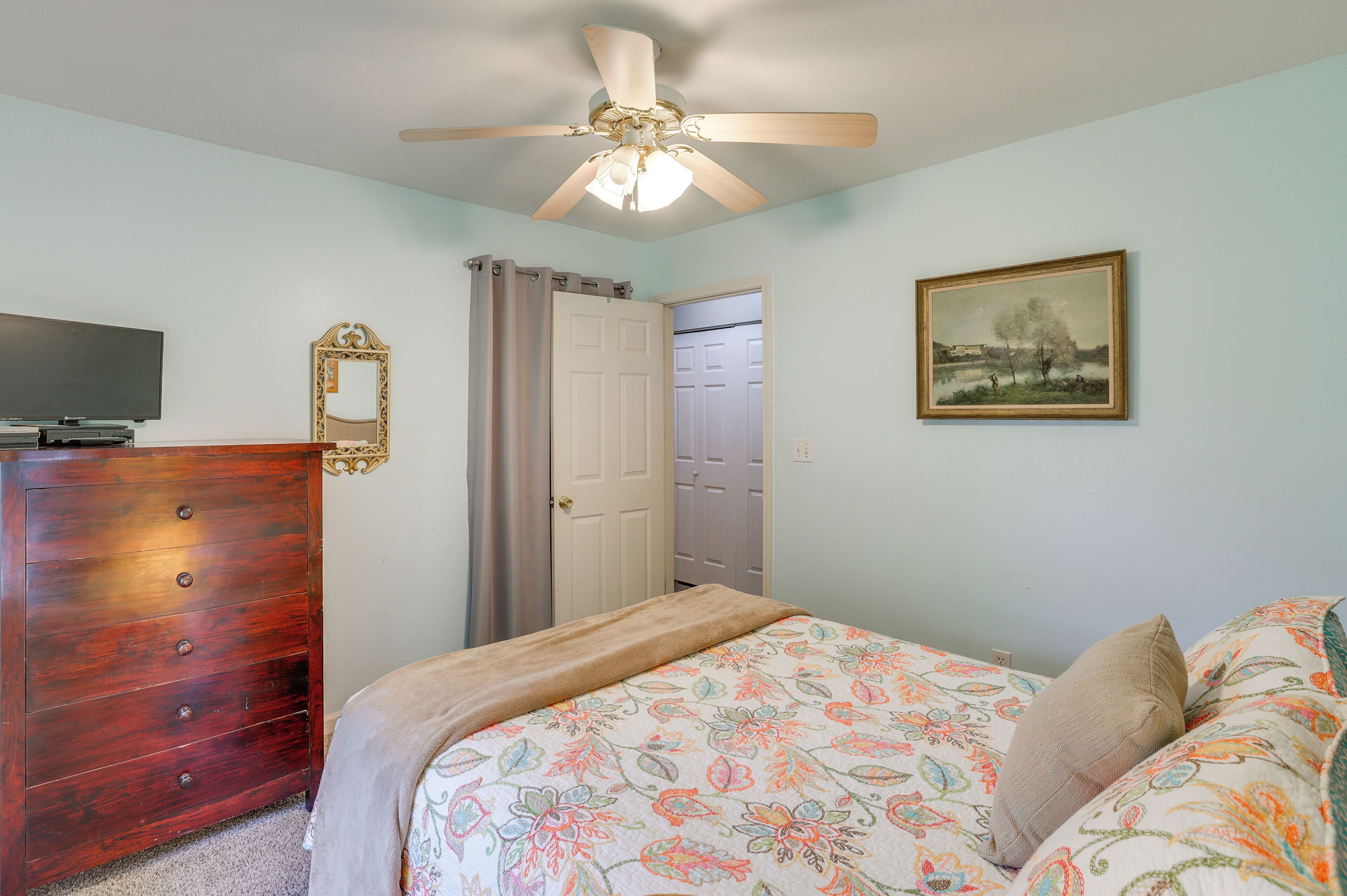 A bedroom with a wooden dresser, a ceiling fan, a bed with floral bedding, a painting, and a door leading to another room.