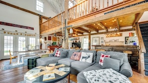 A spacious living room with a large gray sectional sofa, a round coffee table with papers, and a wooden staircase leading to an upper level. - True North Farm House - Historic Family Home (Irasburg)