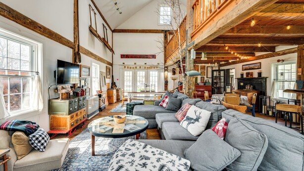 A spacious living room with a large gray sectional sofa, a round coffee table, and a fireplace. There are wooden beams on the ceiling, and a television is mounted on the wall. - True North Farm House - Historic Family Home (Irasburg)