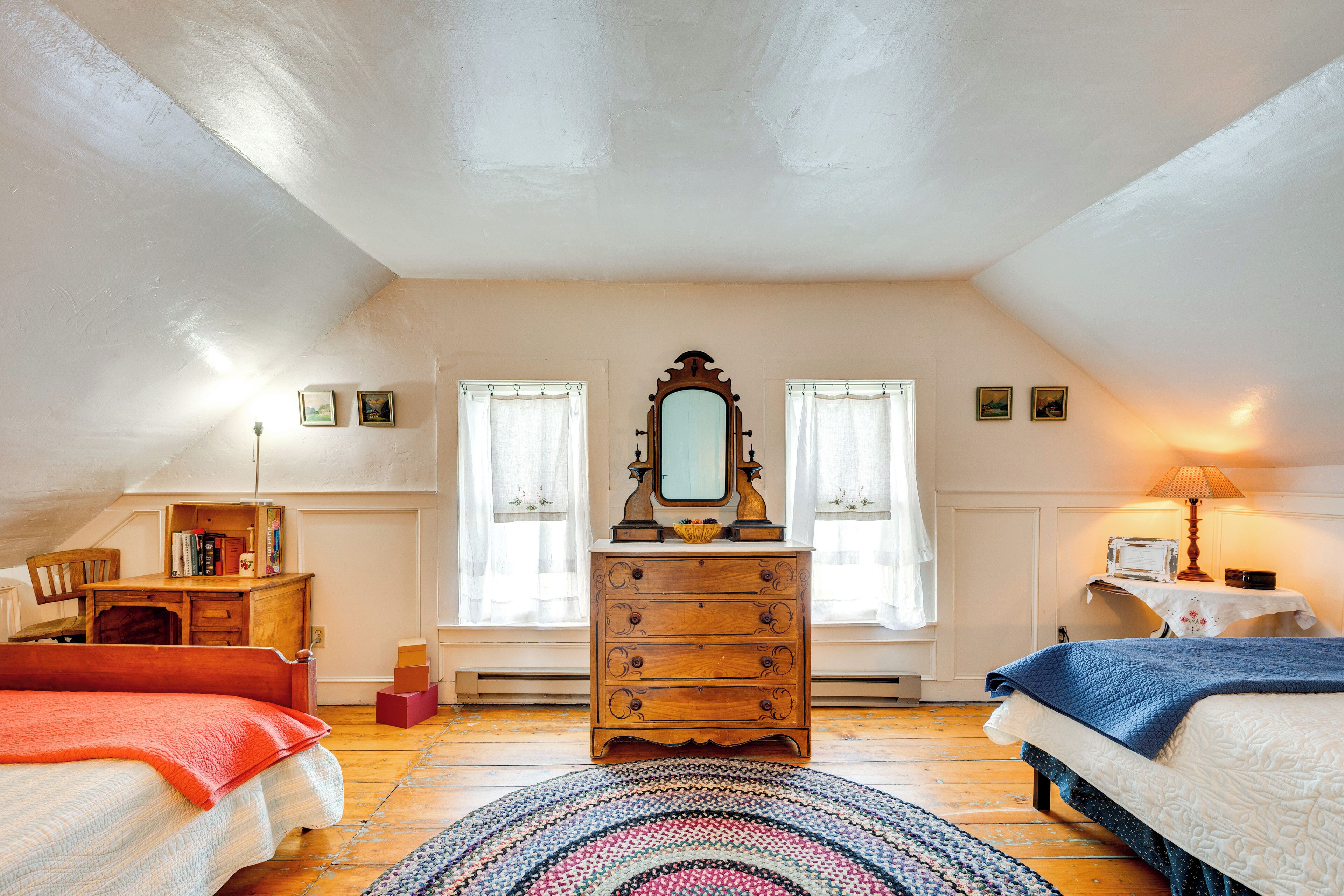 Une chambre avec une commode en bois, un lit recouvert d’une couverture bleue, un tapis rond et une fenêtre avec des rideaux.