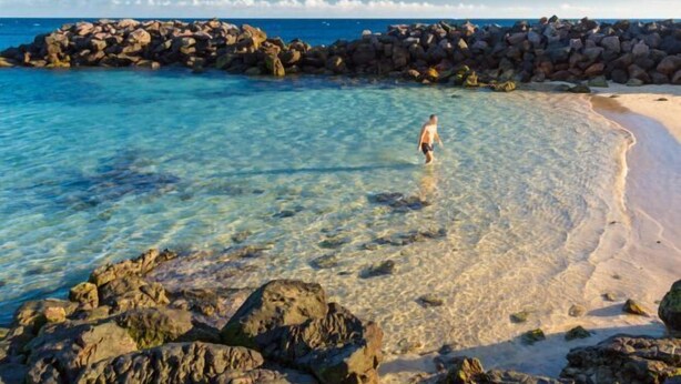 A beach with clear water, a rocky breakwater, and a person standing in the water. - Villa Vista Mar (Tias)