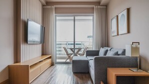 Une chambre d’hôtel moderne avec un canapé gris, un meuble TV en bois, une petite table et un balcon offrant une vue sur la ville.