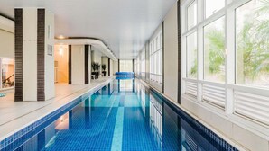 Une longue piscine intérieure aux carreaux bleu clair, bordée d’une rangée de grandes fenêtres d’un côté et d’un mur composé de lattes de bois verticales de l’autre.