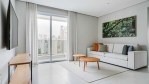 A modern living room with a large window, a flat-screen TV, a white sofa, and a painting of a forest.