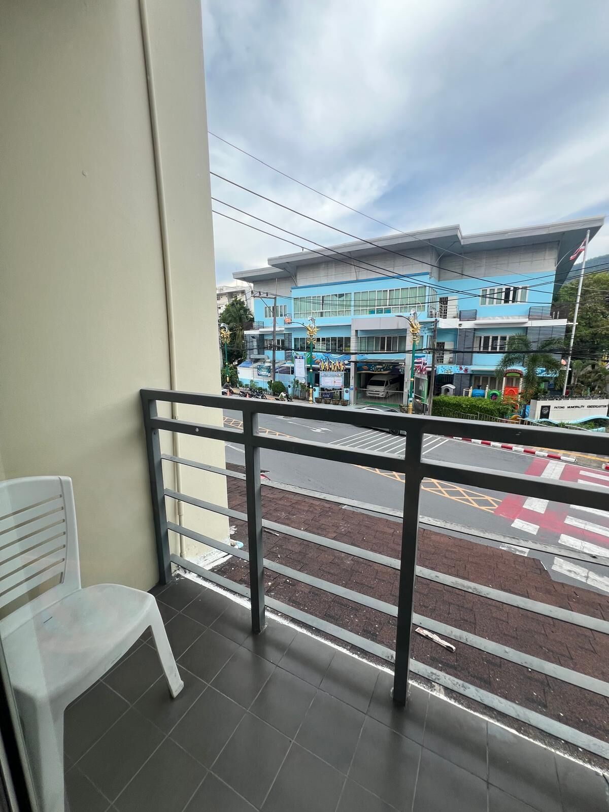 A balcony with a white plastic chair, a metal railing, and a view of a blue building with a parking lot in the foreground.