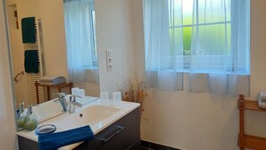 A bathroom with a large mirror, a white sink, a towel rack with towels, a window with sheer curtains, and a vase with dried plants.