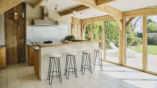 Une cuisine avec des poutres en bois, un grand îlot central doté de tabourets et une vue sur le jardin.