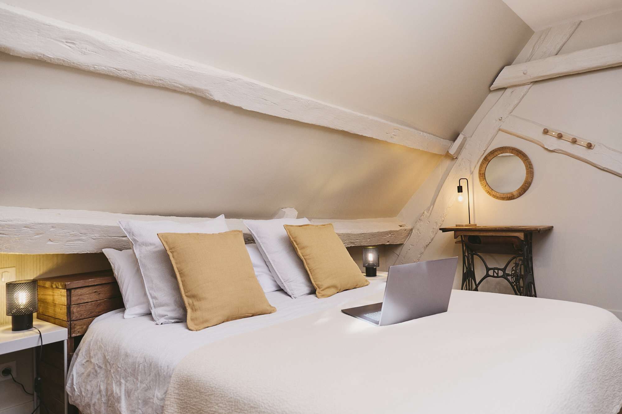 Ein Schlafzimmer mit einem Bett, Kissen, einem Laptop, einer Nachttischlampe, einem kleinen Tisch und einem an der Wand befestigten Spiegel.
