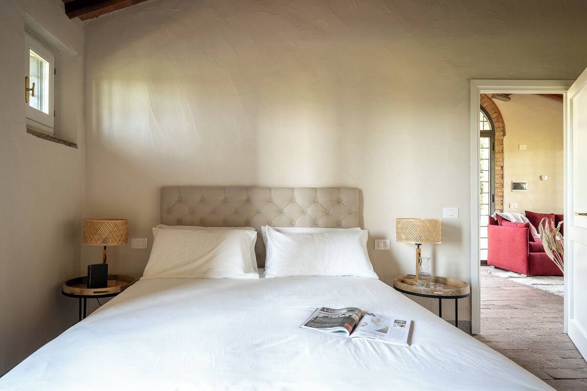 Un dormitorio con una cama grande, dos mesitas de noche con lámparas, una ventana y vistas a un sofá rojo y una pared de ladrillo en la habitación contigua.