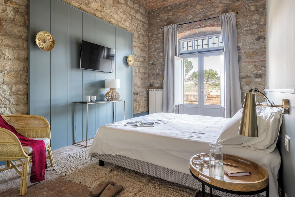 Un dormitorio con cama, televisor, ventana con cortinas, una silla de mimbre y una mesita con lámpara y una botella.