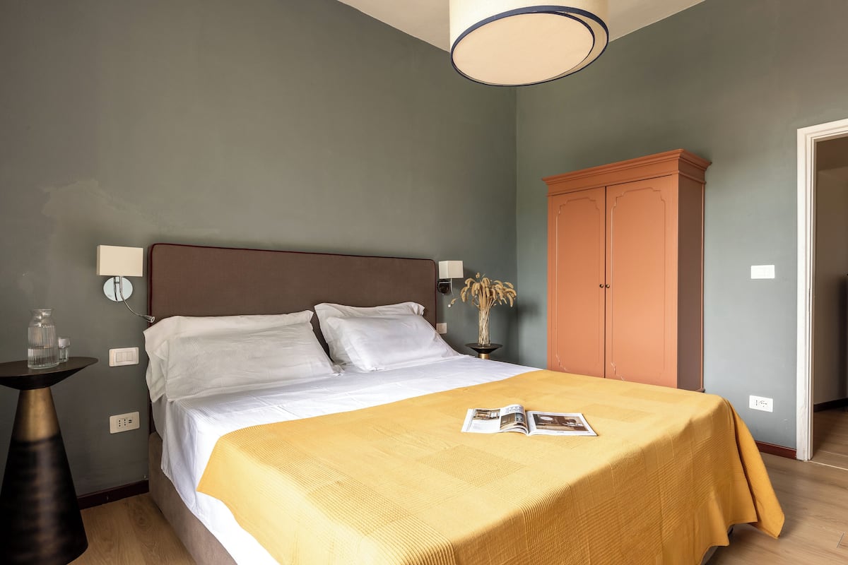 Una cama bien hecha con una colcha amarilla, una mesita de noche con una lámpara y un armario en una habitación con paredes grises.