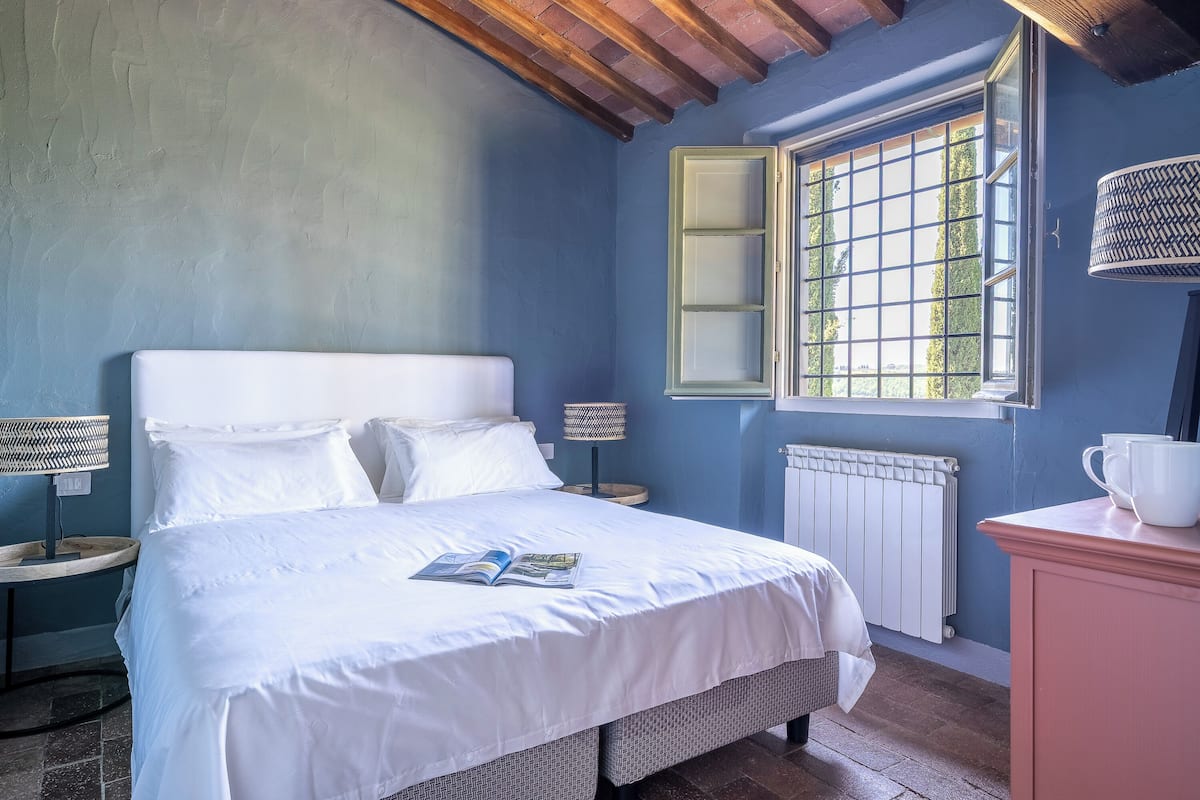 Un dormitorio con cama, ventana con contraventanas, radiador y mesita de noche con lámpara.