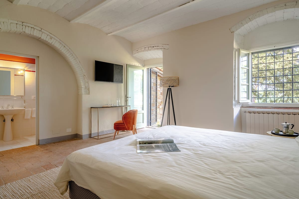 Un dormitorio con cama, escritorio, silla, lámpara y ventana con vista a árboles.